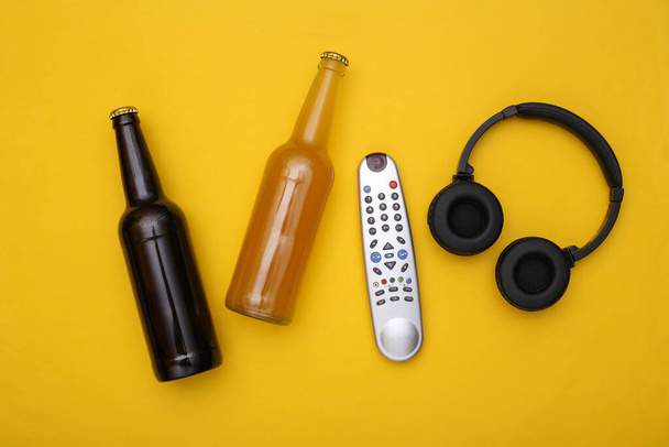 Intrattenimento e tempo libero. Bottiglie di birra, telecomando tv e cuffie stereo su sfondo giallo. Vista dall'alto - Foto, immagini