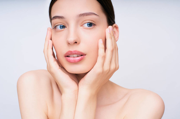 Delicate young girl with bare shoulders, touches her hands face with perfect healthy skin, opened her plump lips and looks to the side. The concept of beauty and care, taking care of the skin. - Photo, image