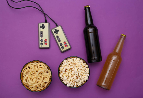 Entertainment time. Gamepads and snack bowl, beer bottles on purple background. Top view - Photo, Image