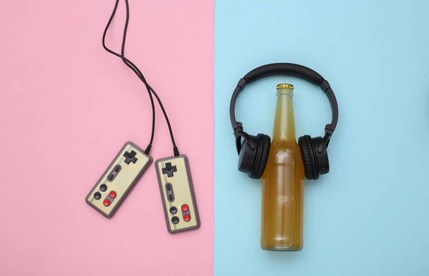 Entertainment time. Gamepads and beer bottle with headphones on pink blue pastel background. Top view - Photo, Image