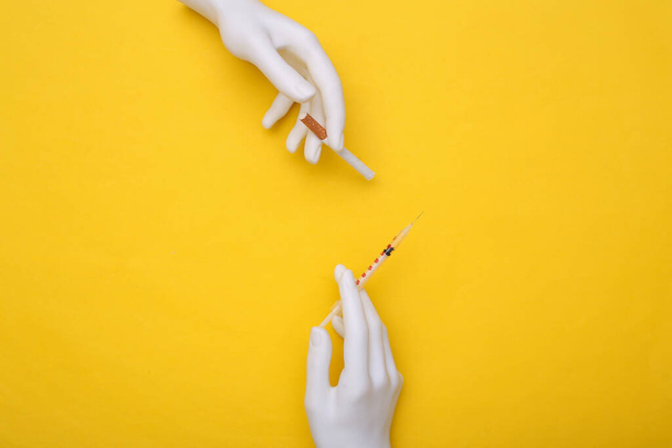 Weiße Hände einer Schaufensterpuppe halten eine Spritze und eine Zigarette auf gelbem Hintergrund. Drogen- und Rauchersucht - Foto, Bild