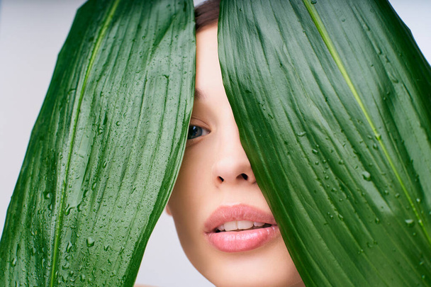 Close-up, a young beautiful girl covers the ldite with green tropical leaves, visible part of the face, women's lips with natural makeup. The concept is natural beauty, naturalness, eco cosmetics. - Photo, image