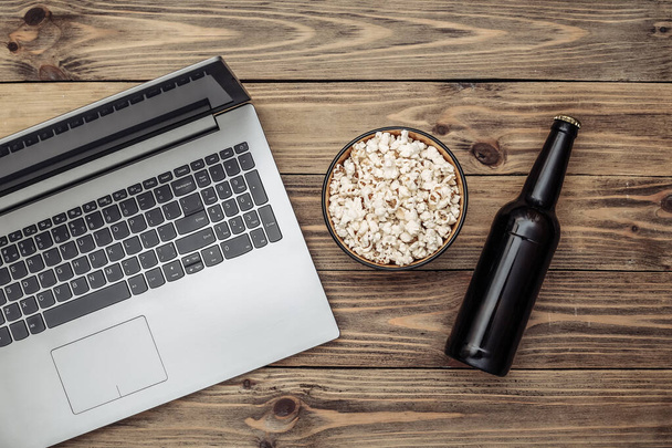 Rest time. Laptop, bottle of beer and popcorn bowl on  wooden background. Top view - Photo, Image