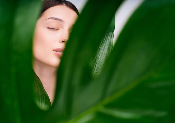 Young beauty brown-haired model with natural make-up closed her eyes, enjoying the beauty, through the green leaf. Spa and wellness. Youth, teenage and skin care concept. Close up, selective focus. - Fotó, kép