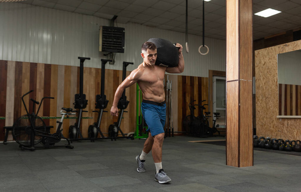 Мускулистый спортсмен мужского пола тренируется с тяжелой сумкой в современном кросс-тренажерном зале. Функциональное обучение - Фото, изображение