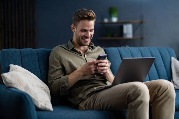Young freelancer using smartphone and smiling. Happy man using mobile phone apps, texting message, browsing internet, looking at smartphone, sitting at home. Young people working with mobile devices - Zdjęcie, obraz