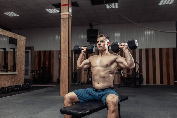 Tehokas urheilullinen mies harjoittelee olkapään lihaksia raskailla käsipainoilla istuessaan penkillä kuntosalilla. Terveiden elämäntapojen käsite - Valokuva, kuva