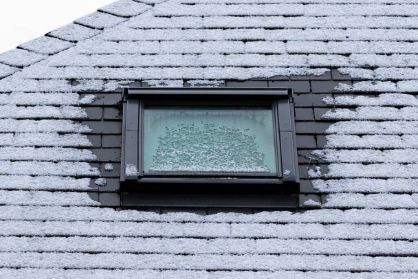 Een portret van een bevroren leistenen dak met een dakraam dat ook 's winters bevroren is. Aan de zijkanten van het raam is de vorst gesmolten door slechte isolatie. - Foto, afbeelding