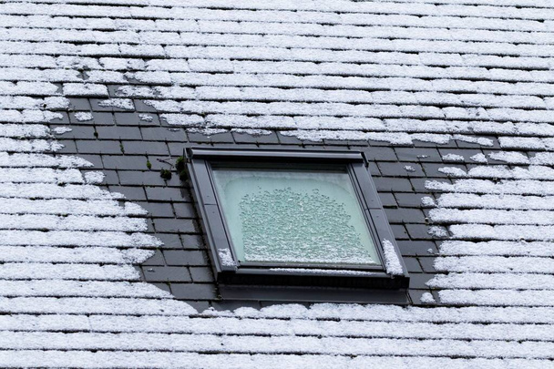 Een verticaal portret van een bevroren leistenen dak met een kantelbaar dakraam dat ook in de winter bevroren is. Aan de zijkanten van het raam is de vorst gesmolten door slechte isolatie. - Foto, afbeelding