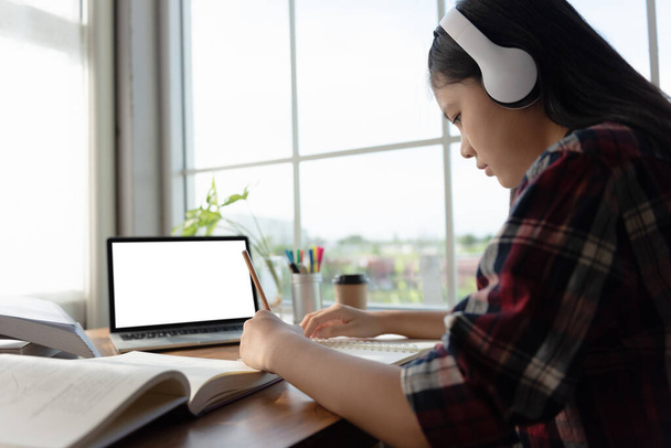 Female student wears headphones sitting at table to learning and note, using laptop for homework during covid19 pandemic and lock down, Social Distance, New life of Student on internet. Mockup screen. - Photo, Image