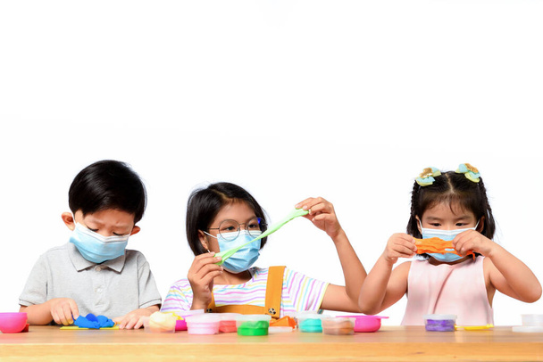 Aziatische kinderen dragen gezichtsmasker tijdens het spelen samen met klei speelgoed op een houten tafel met witte achtergrond. Nieuw normaal voor kinderen, Coronavirus bescherming voor kinderen, COVID symptomen bij kinderen. - Foto, afbeelding