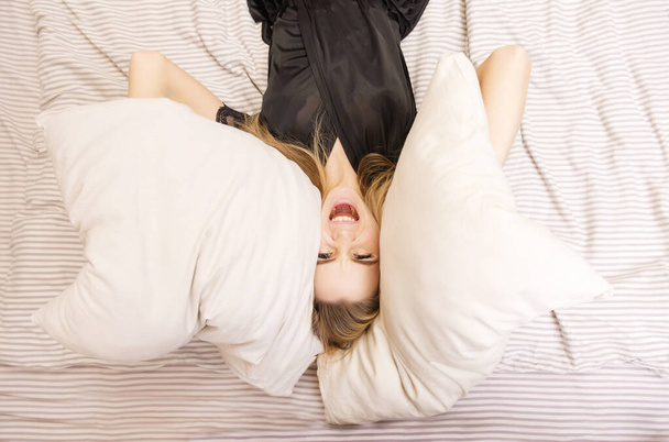 1 femme blonde blanche en peignoir noir noir couchée au lit et tenant des oreillers et criant, souriant, - Photo, image