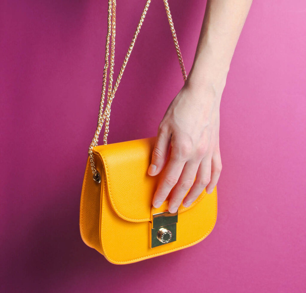 Female hand hold and opens fashionable yellow leather bag with golden chain on purple background. - Photo, Image