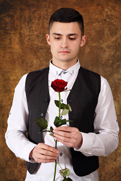 Όμορφος σκεπτικός τύπος με μαύρο γιλέκο και τριαντάφυλλο στα χέρια του Αγίου Βαλεντίνου. - Φωτογραφία, εικόνα