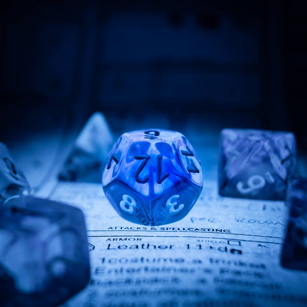 Прозрачный полимерный кубик D12 освещенный снизу мягким голубым светом на подземелье и листе персонажей драконов.  - Фото, изображение
