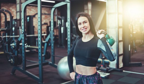 Smilling brunette raises kettlebell with one hand and looking at the camera in the gym. Free-weight strength training, functional training - Photo, Image