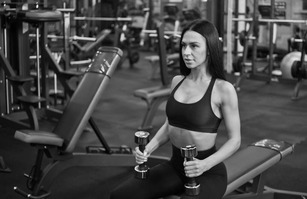 Mujer joven concentrada en el entrenamiento sostiene pesas en las manos mientras está sentada en un banco en el gimnasio - Foto, imagen