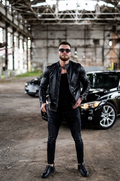 Handsome young man in leather jacket holding a gun. Cool sunglasses on brutal man. - Photo, image