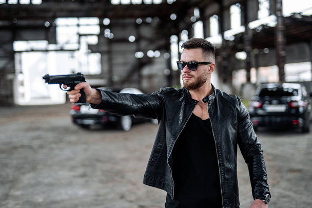 Brutal male points a gun on a point. Man dressed in leather black jacket and fashion glasses. Broken building with no windows on a background. - Zdjęcie, obraz