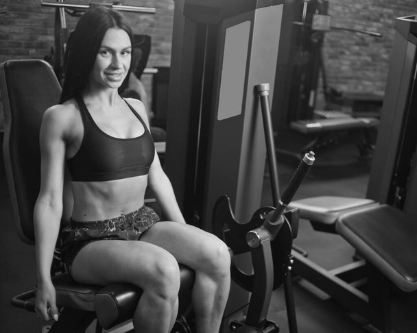 Atrakcyjna brunetka w odzieży sportowej siedząca w maszynie do ćwiczeń i patrząca w kamerę - Zdjęcie, obraz