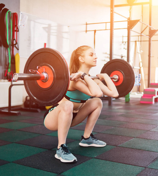Fiatal atléta nő sportruházatban csinál elöl guggoló egy súlyzóval a mellkasán a tornateremben - Fotó, kép