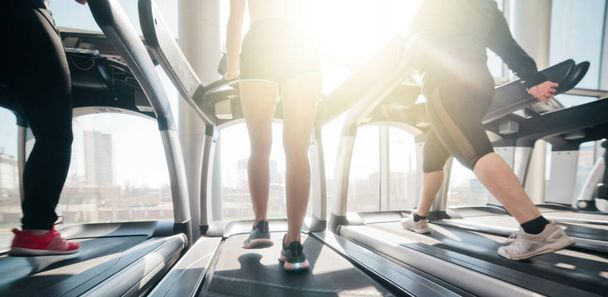 Foto recortada de piernas femeninas en ropa deportiva y zapatillas de deporte corriendo en una cinta de correr en el gimnasio contra la ventana - Foto, imagen