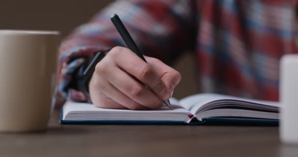 Zbliżenie ujęcia człowieka piszącego plan w notatniku lub wypełniającym dzienniku, siedzącego przy biurku z kawą w domu - Materiał filmowy, wideo