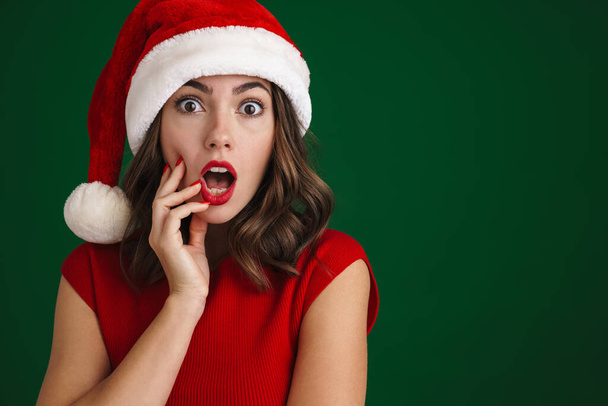 Beautiful shocked girl in Santa Claus hat expressing surprise at camera isolated over green background - Fotografie, Obrázek