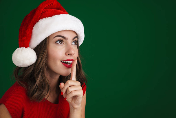 Beautiful excited girl in Santa Claus hat showing silence gesture on camera isolated over green background - Fotó, kép