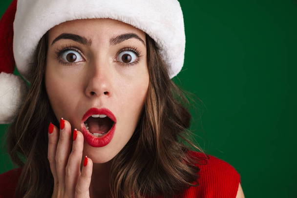 Beautiful shocked girl in Santa Claus hat expressing surprise at camera isolated over green background - Foto, Bild