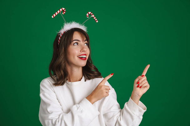 Beautiful happy girl wearing toy horns smiling and pointing fingers aside isolated over green background - Foto, imagen