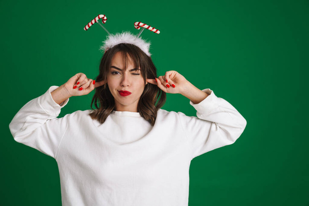 Beautiful displeased girl wearing toy horns winking and plugging her ears isolated over green background - Foto, imagen