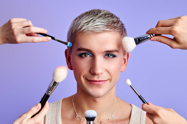 young caucasian transvestite man among lots of brushes pointing at his face - Photo, Image