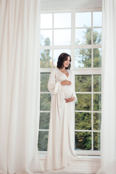 όμορφη γυναίκα που ποζάρει κοντά στο παράθυρο. Ευτυχισμένη έγκυος γυναίκα φορώντας μακρύ λευκό κομψό φόρεμα. Γυναίκα με φόρεμα διακοπών. πορτρέτο μόδας - Φωτογραφία, εικόνα