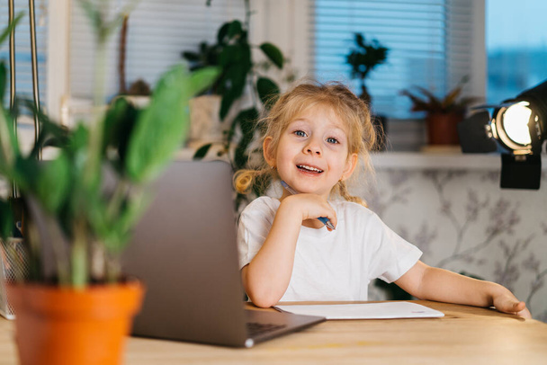 Onnellinen pieni tyttö vlogger istuu kannettavan tietokoneen katselee kameraa, hymyilee ja tekee online video puhelun blogi tallennus, muotokuva - Valokuva, kuva