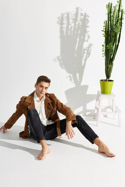 A handsome young man sits on the floor in a jacket and black trousers and a flower in a pot on a chair - Фото, изображение