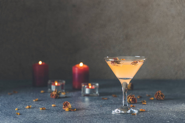 Літній коктейль з грейпфрута мартіні з сушеними трояндами квітами та пелюстками, оточені свічками на темно-сірій поверхні столу
. - Фото, зображення