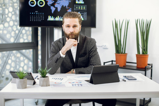 Handsome purposeful confident bearded businessman, chief executive officer in formal wear, sitting at workplace and looking at camera. Male worker sitting at the table in office - Fotoğraf, Görsel
