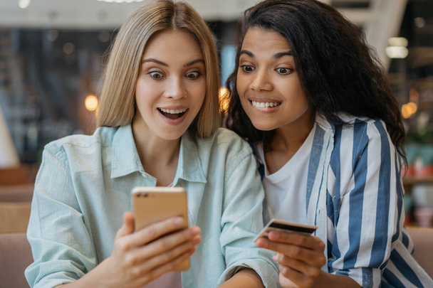 Happy emotional friends holding credit card, using mobile phone, shopping online at home. Young beautiful women ordering food, delivery concept   - Фото, зображення