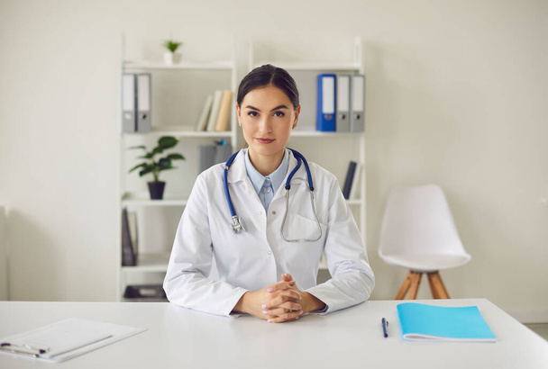 Young general practitioner looking at camera while sitting at table in her office - Foto, imagen