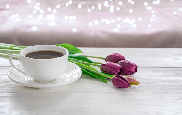 Una taza de café y tulipanes sobre un fondo claro con luces ardientes. - Foto, Imagen
