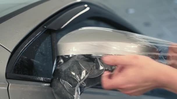 Le mani dei lavoratori avvolgono il film bagnato di protezione della vernice o il rivestimento antighiaia sullo specchio dell'automobile. Dettagli dell'auto - Filmati, video