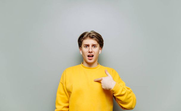 Jeune homme en vêtements jaunes et écouteurs sans fil se tient sur un fond gris et pointe du doigt lui-même avec un visage surpris. - Photo, image