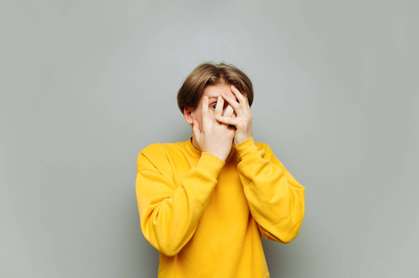 Страшний молодий чоловік в жовтому одязі стоїть на сірому тлі, ховаючись руками на обличчі і заглядаючи в камео. Портрет наляканого хлопця
. - Фото, зображення