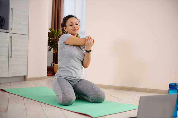Joven mujer deportiva sentada en una alfombra de fitness y estirando su cuerpo a la izquierda mientras hace ejercicio en casa - Foto, imagen
