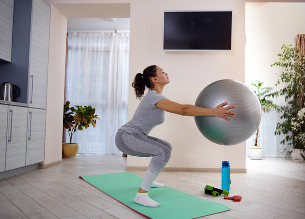 Boční portrét sportovní ženy držící fitness míček a dřepící na podložce na jógu. Cvičení doma během karantény - Fotografie, Obrázek