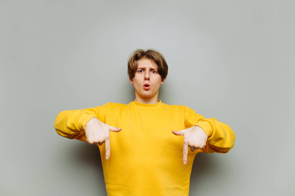 Surprised guy in a gray sweatshirt stands on a background of a gray wall and shows his fingers down on the copy space. - Photo, Image