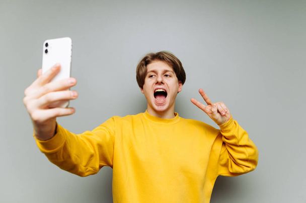 Happy guy in yellow sweatshirt takes selfie on smartphone and shows peace gesture to camera isolated on gray background. - Foto, Imagen