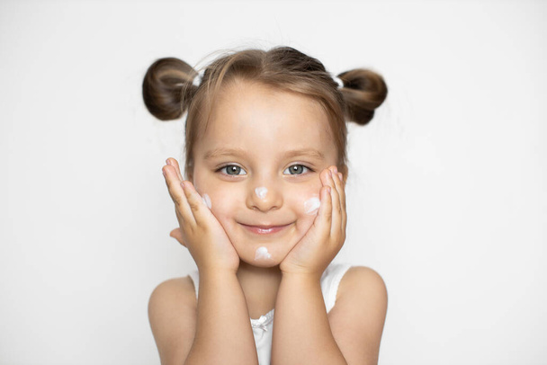 Child face skin care and protection, cold cream. Portrait of cute little caucasian 3 years old girl, applying winter cream, posing on white studio background and smiling - 写真・画像