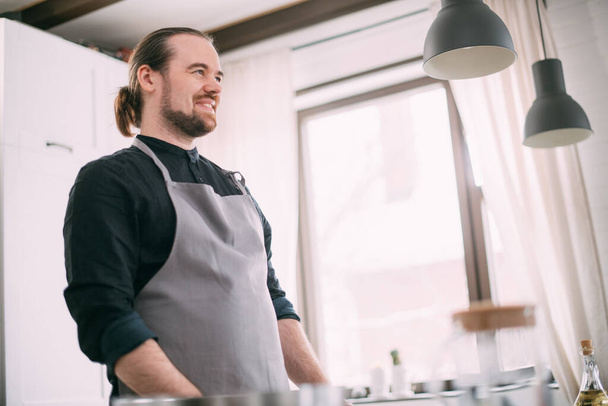 Un cuisinier masculin cuisine à la maison dans la cuisine. Jeune homme dans un tablier dans sa cuisine à la maison à la table avec des ingrédients pour cuisiner le dîner - Photo, image